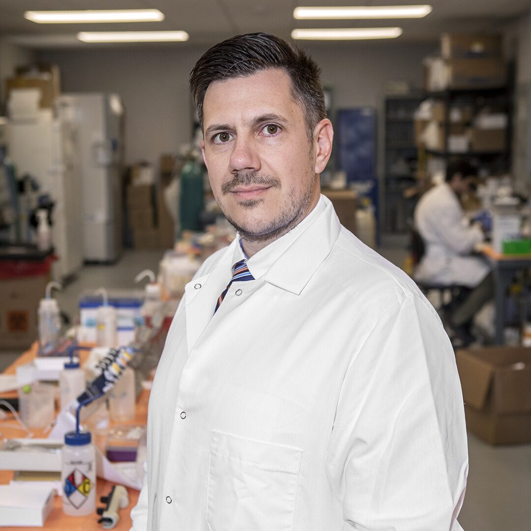 A person in a white lab coat stands in a laboratory, surrounded by equipment and supplies, with another individual working in the background.